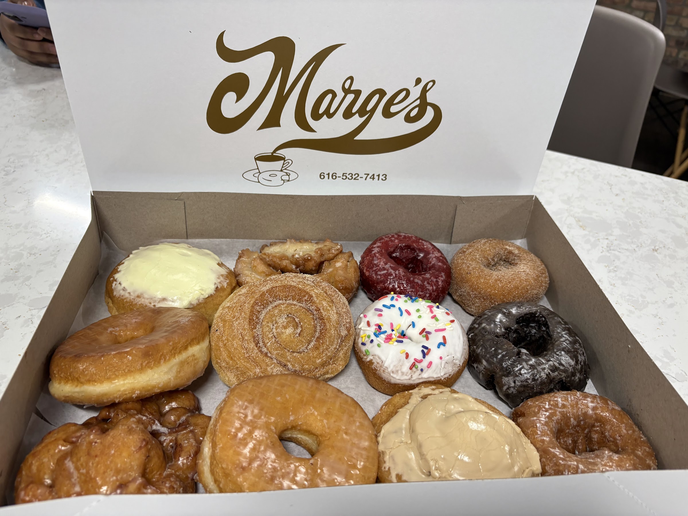 An open box of Marge's mixed dozen — apple fritter, custard long john, cinnamon roll, maple-glazed and sprinkled cake donuts — under the gold cursive Marge's lid